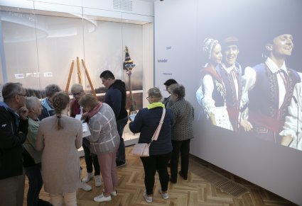Grupa ludzi na ekspozycji w Pałacu Karolin, zwiedzają salę poświęconą pomorzu, na ścianie wyświetlany film Mazowsza w kostiumach ludowych, w gablotach regionalne instrumenty