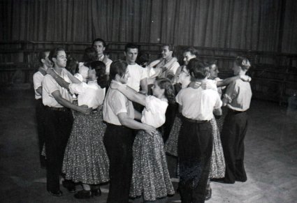 Czarno białe zdjęcie, na parkiecie w kole stoi grupa tancerzy. Mężczyźni na przeciwko kobiet w parach.  Kobiety trzymają ręce na szyjach mężczyzn, mężczyźni trzymają kobiety w pasie