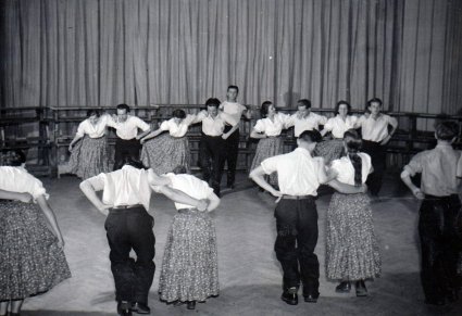 Czarno białe zdjęcie, na parkiecie scenicznym grupa tancerzy. Tancerze stoją w parach,  pochylają się do środka, mężczyźni stoją po lewej stronie kobiet, trzymają je w pasie