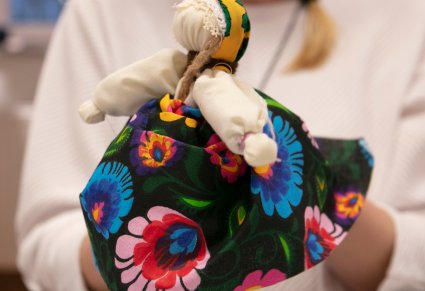 Kobieta trzyma materiałową lalkę w dłoniach, lalka ubrana w kolorowy materiał, spódnica we wzory łowickich kwiatów.