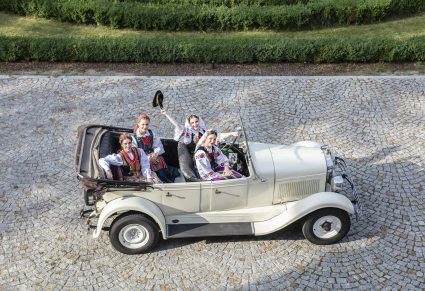 Zdjęcie wykonane z góry, w zabytkowym samochodzie zaparkowanym na kostce siedzą cztery młode kobiety, uśmiechają się do zdjęcia, ubrane są w kostiumy ludowe