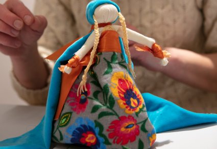 Lalka motanka ubrana w kolorowy kostium, sukienka ze wzorem łowickich kwiatów, lalka na głowie ma warkoczyki i niebieską, długą chustę.