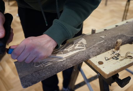 Zbliżenie: Na zdjęciu uczestnik warsztatów w dłoni trzyma dłuto, którym rzeźbi wzory w podłużnym kawałku drewna