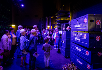 Goście na zwiedzaniu zakulisowym podczas Nocy Muzeów 2024 oglądają przestrzeń za sceną na Sali Widowiskowej Zespołu "Mazowsze". Przewodnik opowiada, wskazując na skrzynie w których kostiumy są przechowywane