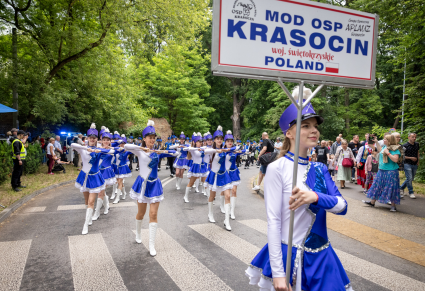 Przemarsz Młodzieżowej Orkiestry Dętej OSP Krasocin. Na przodzie mażoretki idą ulicą, po bokach ogląda ich publiczność