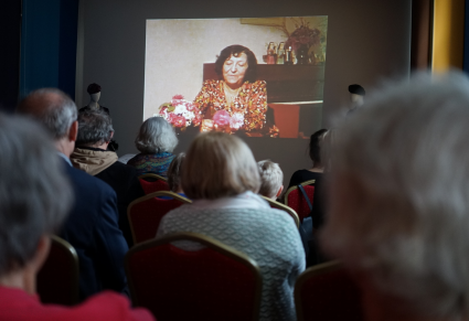 Uczestnicy spotkania siedzą na krzesłach w Sali Rekreacyjnej Centrum Folkloru Polskiego "Karolin", patrząc w stronę projektora na którym wyświetlone jest zdjęcie Miry Zimińskiej-Sygietyńskiej w Karolinie