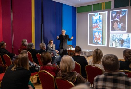 Profesor Pokropek przemawia do publiczności przy projektorze na którym widać zdjęcia artystów ludowych oraz ich prace.