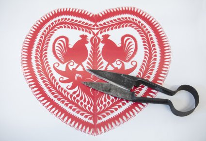 Czerwona wycinanka w kształcie serca, po środku wycięte dwa koguty. Na wycinance leżą stare nożyce do przycinania owiec
