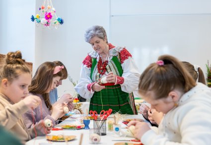 Starsza kobieta stoi przy stole warsztatowym i pokazuje jak okleja jajko, przy stole siedzą uczestnicy warsztatów, w rękach trzymają swoje jajka