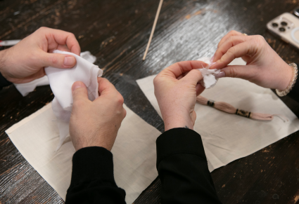 Zbliżenie: dłonie uczestników warsztatów trzymające kawałki złożonego materiału. Na stole leży płótno pocięte w kwadraty