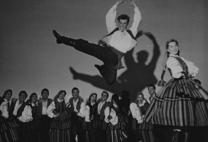 Para tańcząca w strojach opoczyńskich, mężczyzna wyskakuje wysoko unosząc ręce, z tyłu grupa przyglądających się osób