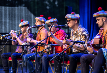 Członkowie Warszawskiego Combo Tanecznego z beretami na głowach siedzą na scenie z instrumentami - gitary, banjo, trąbka, saksofon - w rękach.