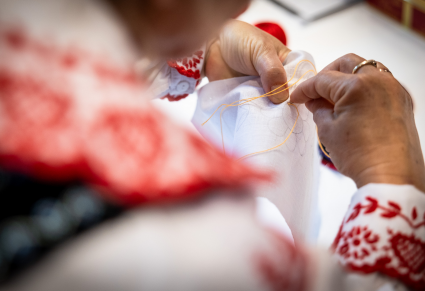 Osoba prowadząca warsztaty w stroju ludowym przeciąga igłę z nitką przez materiał podczas warsztatów z haftu lachowskiego.