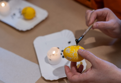 Uczestnik warsztatów w jednym ręku trzyma dekorowane jajko barwione na żółto, a w drugim lejek do wosku. Na stole przed nim świeczka na papierowym talerzyku