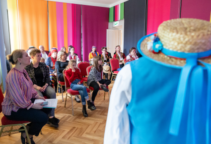 Uczestnicy spotkania siedzą na krzesłach w Sali Rekreacyjnej Centrum Folkloru Polskiego. W rogu zdjęcia na pierwszym planie manekin z kapeluszem słomianym z niebieską tasiemką, ubrany w białą koszulą i niebieską kamizelką