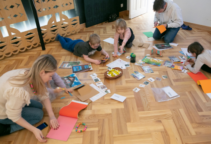 Grupa dzieci i dorosłych siedzących na podłodze, przy nich kolorowy papier, klej, nożyce i zdjęcia domów z malowanymi okiennicami. Dzieci i dorośli tną, kleją i rysują