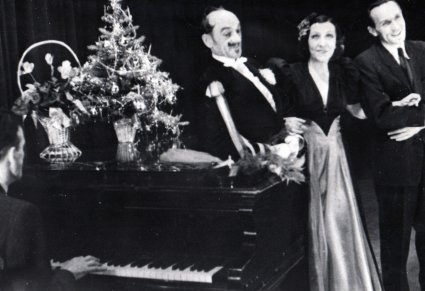 Czarno białe zdjęcie, po lewej fortepian przy którym gra mężczyzna na fortepianie stoi choinka i kwiaty, po prawej Mira z dwoma mężczyznami, uśmiechnięci odgrywają role teatralne.