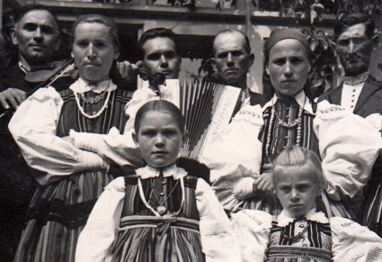 Grupa ludzi ubrana w stroje opoczyńskie, z przodu stoją dwie dziewczynki, za nimi dwie kobiety, z tyłu mężczyźni z harmoniami i skrzypcami w rękach.