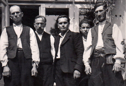 Czarno białe zdjęcie na nim pięciu mężczyzn, dwóch z nich ubranych w kostiumy ludowe, inni ubrani w garnitury, pozują do zdjęcia na tle okien