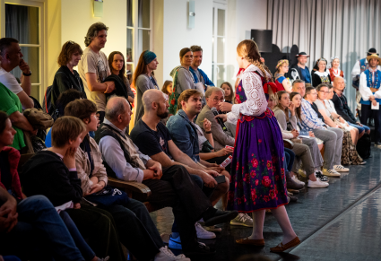 Pracowniczka Centrum Folkloru Polskiego "Karolin" w kostiumie podhalańskim rozmawia z członkami widowni podczas wydarzenia "Pokaz Mazowszańskiej Mody"