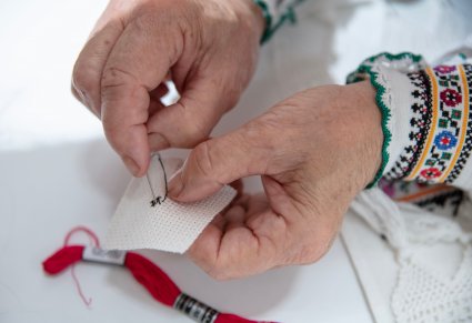 Twórczyni ludowa pokazuje haft krzyżykowy, przewlekając igłę z nitką przez materiał. Zdjęcia pokazuje dłonie w zbliżeniu