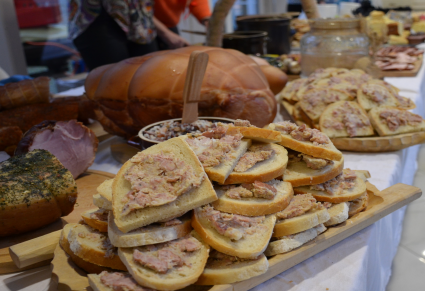 Pokrojone kromki chleba posmarowane smalcem leżą na desce na stole.  W tle duża szynka, różne rodzaje mięs, słoik i miski