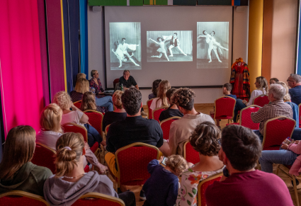 Uczestnicy spotkania siedzący na krzesłach w Sali Rekreacyjnej Centrum Folkloru Polskiego, patrzą w strone Michała Jarczyka, za którym wyświetlone na projektorze są zdjęcia jego w tańcu klasycznym