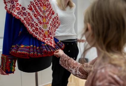 Młoda uczestniczka warsztatów z bliska ogląda haftowaną chustę oraz kaftan z organikami - elementy kostiumu podegrodzkiego Zespołu "Mazowsze"