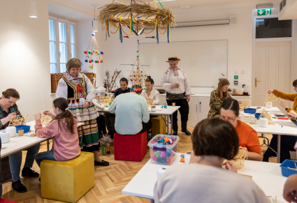 Uczestnicy warsztatów siedzą przy stołach na sali edukacyjnej Centrum Folkloru Polskiego, pochyleni nad konstrukcjami ze słomy. Przy suficie wiszą pająki a przy nich stoi para prowadzących strojach krzczonowskich