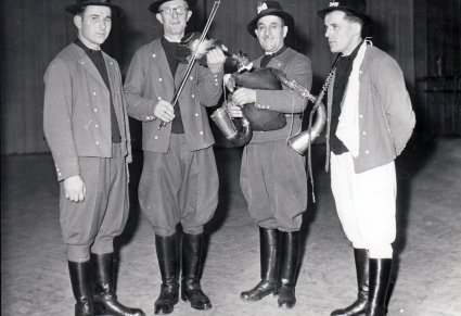 Czterech mężczyzn, dwóch w środku trzyma instrumenty ludowe, skrzypce oraz dudy.