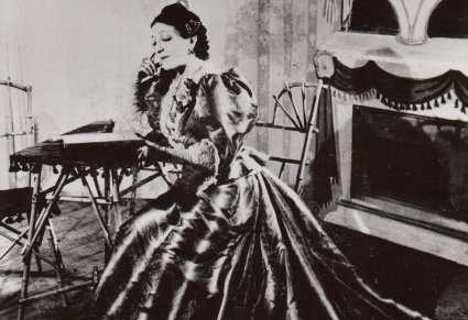 Czarno białe zdjęcie, Mira siedzi przy stoliku, podpiera głowę na prawej ręce, zamyślona, ubrana w suknię, w tle scenografia teatralna.