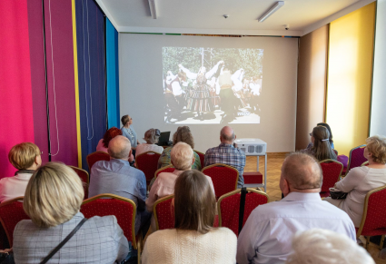 Uczestnicy spotkania siedzą na krzesłach w Sali Rekreacyjnej Centrum Folkloru Polskiego. Przed nimi na projektorze wyświetlone jest zdjęcie czarno-białe archiwalne PZLPiT MAzowsze. Na froncie siedzi Wioletta Milczuk, która prowadzi spotkanie