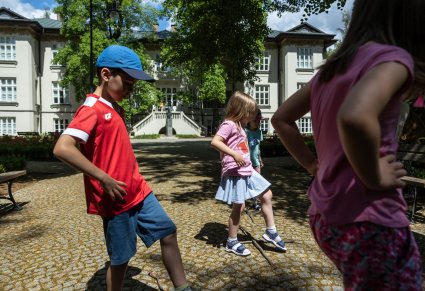Na dworze, w słoneczny dzień, przed Pałacem Karolin, dzieci ćwiczą zabawę przeskakiwania przez skrzyżowane patyki