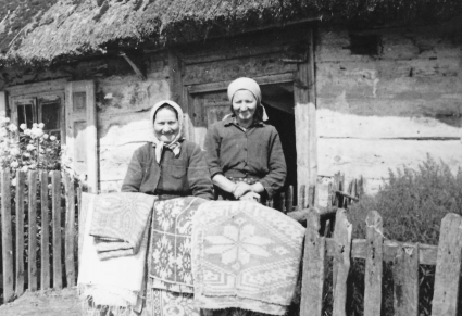 Dwie kobiety stojące przy drewnianym płocie na tle chaty, na płocie zwieszone ludowe tkaniny