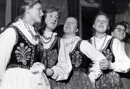Cztery kobiety ubrane w stroje ludowe, śpiewają trzymając się za ręce.