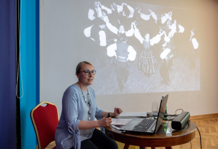 Wioletta Milczuk, osoba prowadząca spotkanie, siedzi przy stole na Sali Rekreacyjnej Centrum Folkloru Polskiego. W tle projektor, na którym jest wyświetlone czarno-białe archiwalne zdjęcie PZLPiT Mazowsze na scenie