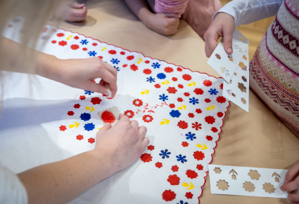 Osoba prowadząca warsztaty pokazuje jak zdobić chustę we wzory kwieciste za pomocą farby w tubce, gąbki i szablonów