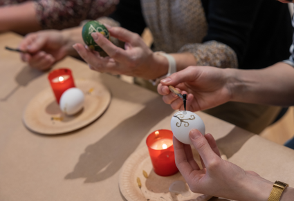 Uczestnicy warsztatów siedzą przy stole, trzymając w rękach jajka udekorowane techniką tak zwaną batikową. Przed nimi na papierowych talerzykach stoją świece.  Jedna z uczestniczek lejkiem nakłada wosk na jajko