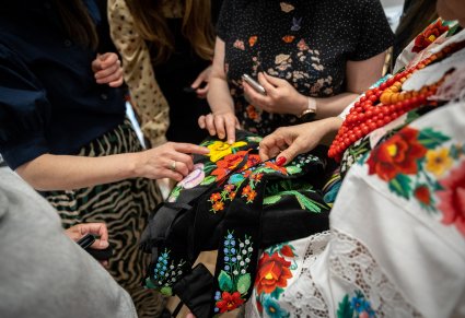 Zbliżenie na dłonie kobiet, jedna z nich ubrana w kostium łowicki trzyma wyhaftowany fragment kostiumu, reszta kobiet dłońmi dotyka do haftu