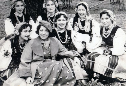 Czarno białe zdjęcie, Mira Zimińska w otoczeniu swoich uczennic z Zespołu, kobiety ubrane w stroje ludowe, Mira w sukience, płaszczu i berecie na głowie, siedzą na trawie w parku.