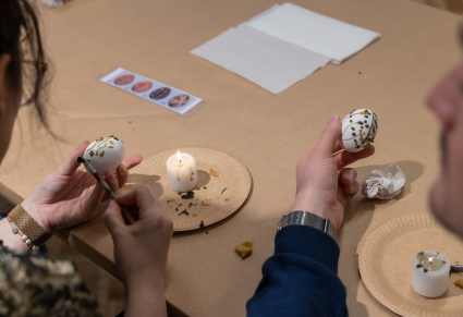 Uczestnicy warsztatów siedzą przy stole, trzymając w rękach jajka udekorowane woskiem. Przed nimi na papierowych talerzykach stoją świece.  Jedna z uczestników trzyma także lejek.