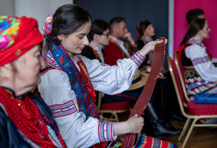Uczestnicy spotkania siedzący na krzesłach w Sali Rekreacyjnej Centrum Folkloru Polskiego, ubrani w strojach krzczonowskich, oglądają pas w kolorze bodowym