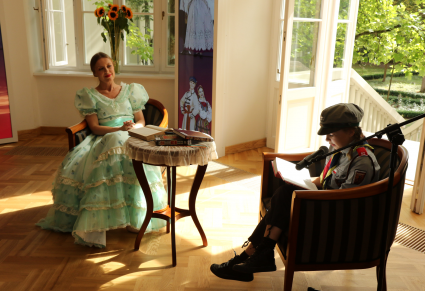 Harcerka siedząca w foyer Pałacu Karolin czyta lekturę "Nad Niemnem". Przy niej mikrofon na statywie. Na przeciwko siedzi przewodniczka CFP w kostiumie stylowym. Pomiędzy nimi stoi stolik.