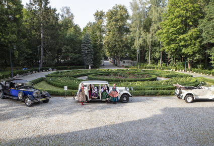 Park w słoneczny dzień, na kostce ustawione są trzy zabytkowe samochody, w środkowym aucie grupa artystów zespołu Mazowsze