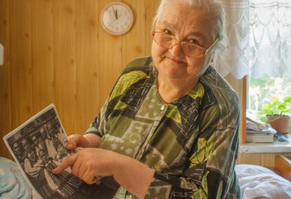 Do zdjęcia pozuje starsza, uśmiechnięta kobieta, w prawej ręce trzyma czarno białe zdjęcie, lewą ręką wskazuje na postać na tym zdjęciu