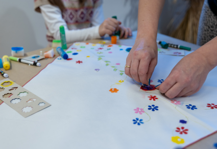 Dwie uczestniczki warsztatów za pomocą szablonu i farby w tubce dekorują chustę wzorami kwiecistymi