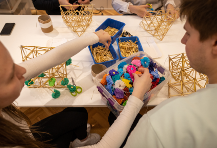 Uczestniczka warsztatów sięga po kawałki słomy i kolorowe kwiatki z bibuły w pojemnikach na stole na którym także leża słomiane konstrukcje