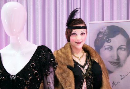 Modelka w czarnej sukience, etoli zfutra i opasce z piórem stoi pomiędzy manekinem ubranym w suknie Miry Zimińskiej a portretem Miry z okresu międzywojennego