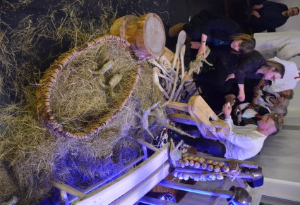 Uczestnicy potańcówki przyglądają się mężczyźnie z drewnianą łyżką w ręku. Na podłodze przed nimi duże drewniane łyżki., koszyk z sianem, figurki z siana i rozsypane siano