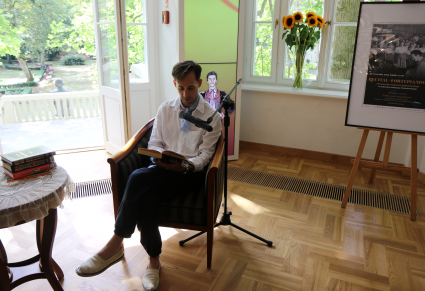 Uczestnik Narodowego Czytania siedzący w foyer Pałacu Karolin czyta lekturę "Nad Niemnem". Przy nim ustawiony mikrofon na statywie.